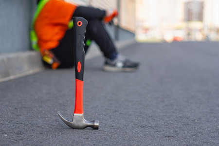 Construction worker in a safety vest rests beside a hammer on an asphalt surface, capturing a moment...