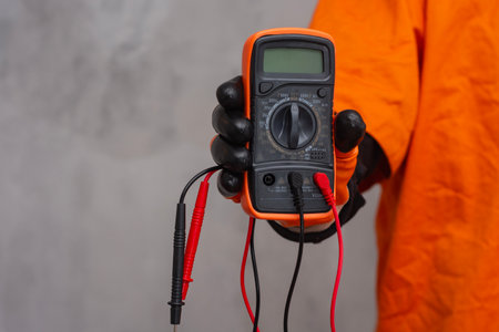 Electrician wearing a protective glove, holding a digital multimeter. Orange colored instrument with connected test leads for measuring voltage, current and resistance in electrical systemsの写真素材