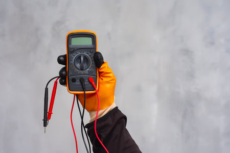 Electrician wearing a protective glove, holding a digital multimeter. Orange colored instrument with connected test leads for measuring voltage, current and resistance in electrical systemsの写真素材