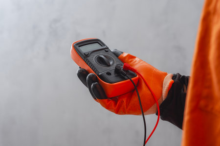 Electrician wearing a protective glove, holding a digital multimeter. Orange colored instrument with connected test leads for measuring voltage, current and resistance in electrical systemsの写真素材