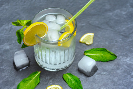 Chilled lemonade glass with ice cubes and green straw. Drink decorated with fresh lemon slices and bright mint leaves on a dark gray textured backgroundの写真素材