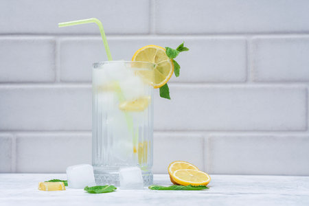 A glass of refreshing lemon water with ice cubes, mint leaves and a straw stands on a light marble background. A citrus drink for a healthy lifestyle, diet and freshnessの写真素材