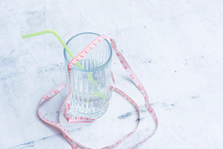 Clear water beverage glass with yellow straw and pink measuring tape on light colored background. Minimalist concept for weight loss, diet and healthy lifestyleの写真素材