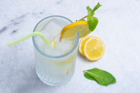 A glass of refreshing lemon water with ice cubes, mint leaves and a straw stands on a light marble background. A citrus drink for a healthy lifestyle, diet and freshnessの写真素材
