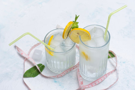 Two glasses of cold diet lemonade with lemon wedges, mint and ice cubes arranged on a marble table with measuring tape. Concept for healthy living, weight loss, detox drinks and fitness nutritionの写真素材