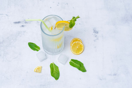 A glass of refreshing lemon water with ice cubes, mint leaves and a straw stands on a light marble background. A citrus drink for a healthy lifestyle, diet and freshnessの写真素材