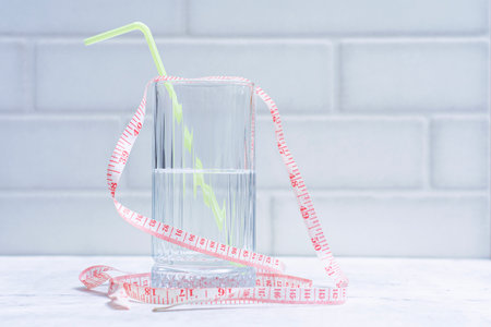 Clear water beverage glass with yellow straw and pink measuring tape on light colored background. Minimalist concept for weight loss, diet and healthy lifestyleの写真素材