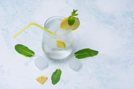 A glass of refreshing lemon water with ice cubes, mint leaves and a straw stands on a light marble background. A citrus drink for a healthy lifestyle, diet and freshnessの写真素材
