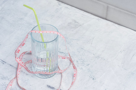 Clear water beverage glass with yellow straw and pink measuring tape on light colored background. Minimalist concept for weight loss, diet and healthy lifestyleの写真素材
