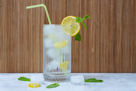 A tall glass of iced lemon water with fresh mint leaves and a green straw, on a light background with ice cubes and lemon slicesの写真素材