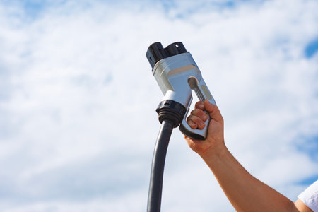 Hand holding an EV charging gun against a bright sky background. Concept sustainable energy and electric vehicle useの写真素材