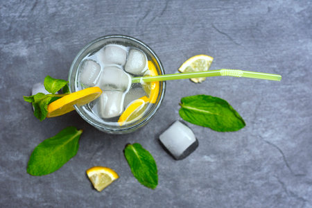 Chilled lemonade glass with ice cubes and green straw. Drink decorated with fresh lemon slices and bright mint leaves on a dark gray textured backgroundの写真素材