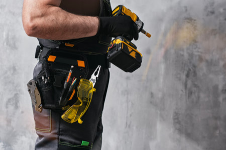 Uniformed construction worker holding a yellow colored cordless screwdriver with a set of tools on his belt. The concept of repair, maintenance and electrical installationの写真素材