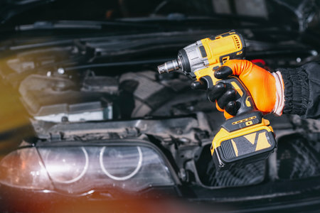 A mechanic wearing protective gloves holds a cordless wrench next to a car engine. Concept with tools and car maintenance in a car repair shopの写真素材