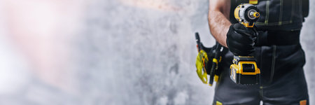 Uniformed construction worker holding a yellow colored cordless screwdriver with a set of tools on his belt. The concept of repair, maintenance and electrical installationの写真素材