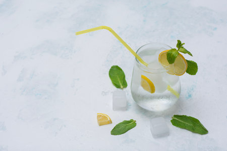 A glass of refreshing lemon water with ice cubes, mint leaves and a straw stands on a light marble background. A citrus drink for a healthy lifestyle, diet and freshnessの写真素材