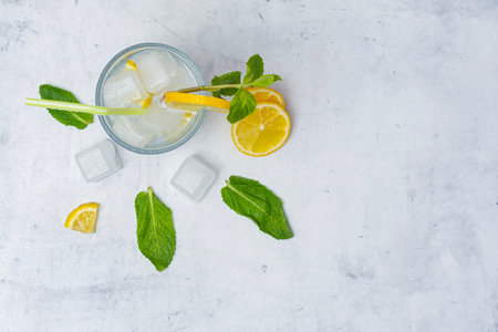 A glass of refreshing lemon water with ice cubes, mint leaves and a straw stands on a light marble background. A citrus drink for a healthy lifestyle, diet and freshnessの写真素材