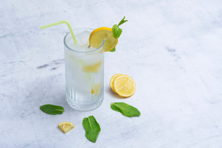 A glass of refreshing lemon water with ice cubes, mint leaves and a straw stands on a light marble background. A citrus drink for a healthy lifestyle, diet and freshnessの写真素材