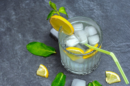 Chilled lemonade glass with ice cubes and green straw. Drink decorated with fresh lemon slices and bright mint leaves on a dark gray textured backgroundの写真素材