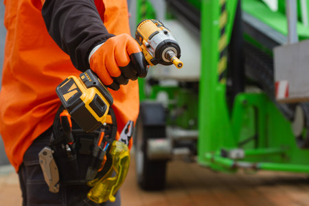 Close-up of a construction worker with a tool belt, wearing protective gloves, holding a cordless drill. Concept on a construction site with green industrial equipmentの写真素材