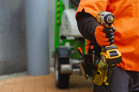 Close-up of a construction worker with a tool belt, wearing protective gloves, holding a cordless...