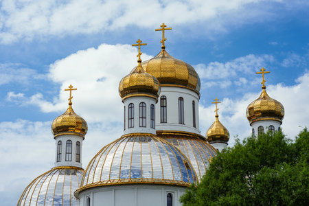 Golden onion domes of an Orthodox church under a blue sky with clouds. Architecture with white walls and gilded crosses. Religious traditions and design of the church in Russian styleの写真素材