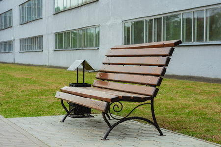 Wooden park bench with a trash can nearby, placed on a paved path beside a modern building with large windows. Peaceful urban setting for outdoor relaxationの写真素材
