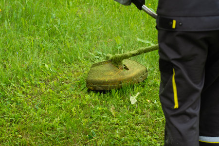 Worker in uniform mowing grass with a string trimmer in a public green area. Manual labor and professional landscaping workの写真素材