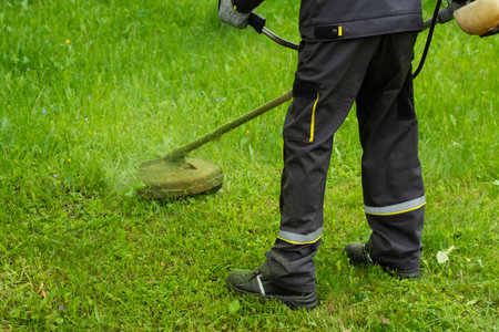 Worker in uniform mowing grass with a string trimmer in a public green area. Manual labor and professional landscaping workの写真素材