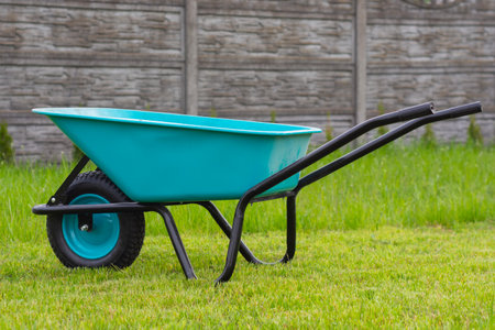 Turquoise colored garden wheelbarrow with one wheel and black metal frame and green grassy lawn with concrete fence in the backgroundの写真素材