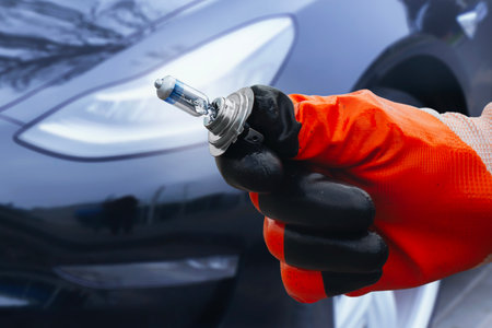 Mechanic wearing protective gloves holds a halogen car headlight bulb near the front of a modern car. Concept of car maintenance or bulb replacementの写真素材