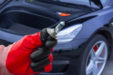 Mechanic wearing protective gloves holds a halogen car headlight bulb near the front of a modern car. Concept of car maintenance or bulb replacementの写真素材