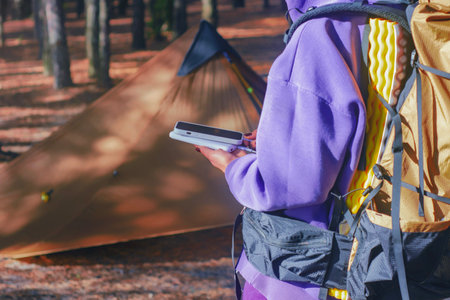 Close-up a woman with a hiking backpack and a smartphone and portable power bank in hands, against the background of a tent in the forest. Digital nomad's concept of sustainable existence in natureの写真素材
