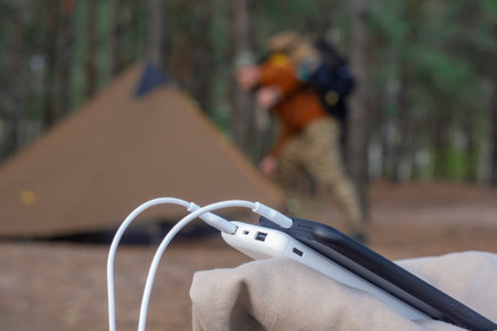 A portable power bank charges smartphones outdoors at a campground surrounded by forest. A hiker with a backpack and a tent in the background. Concept of adventure and digital nomadic lifestyleの写真素材