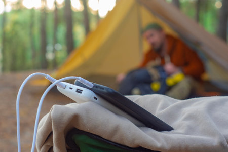 A portable power bank charges smartphones outdoors at a campground surrounded by forest. A hiker with a backpack and a tent in the background. Concept of adventure and digital nomadic lifestyleの写真素材