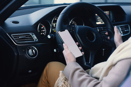 A woman in the driver's seat of a car holding a mockup of a smartphone with a white blank screen. The concept of mobile communication, driving with technology and navigation on the roadの写真素材
