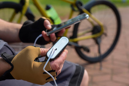 A male cyclist wearing sports gloves charges his smartphone with a portable power bank. The concept of modern technology and active lifestyle in nature, gadgets and energy on the goの写真素材