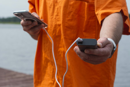 Man charging smartphone with power bank by lake using USB cable in summer outdoor. Concept modern lifestyle, mobile technology, travel, and connectivity on the goの写真素材