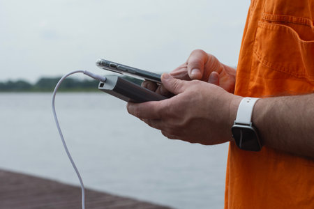 Man charging smartphone with power bank by lake using USB cable in summer outdoor. Concept modern lifestyle, mobile technology, travel, and connectivity on the goの写真素材