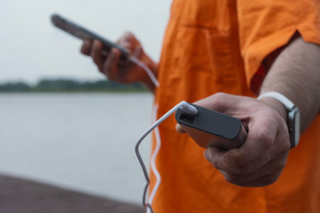 Man charging smartphone with power bank by lake using USB cable in summer outdoor. Concept modern lifestyle, mobile technology, travel, and connectivity on the goの写真素材