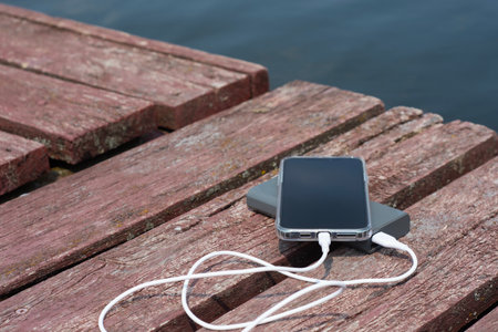 A smartphone connected to a power bank lies on a wooden pier by the water. The concept of travel, autonomy, mobile energy and technology in natureの写真素材