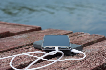 A smartphone connected to a power bank lies on a wooden pier by the water. The concept of travel, autonomy, mobile energy and technology in natureの写真素材