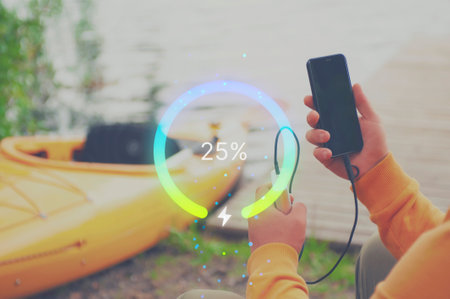 A man is charging a Smartphone with a Power Bank by a lake next to a bright yellow kayak. A glowing battery percentage graph symbolizing real-time charging. Concept on the theme of tourism.の写真素材