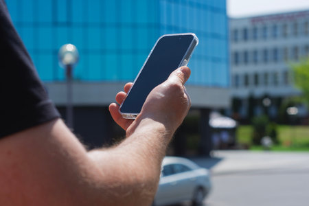 Close-up of a man hand holding a modern smartphone against a mirrored blue glass building in a city. Concept of technological lifestyle in urban environmentの写真素材