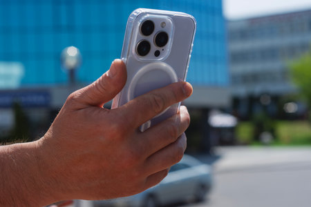 Close-up of a man hand holding a modern smartphone against a mirrored blue glass building in a city. Concept of technological lifestyle in urban environmentの写真素材