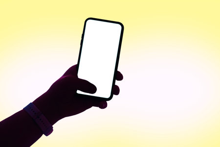 Silhouette of a hand holding a modern smartphone with a blank white screen, isolated on a soft yellow gradient background. Mockup for mobile applications and UI UX design presentationsの写真素材