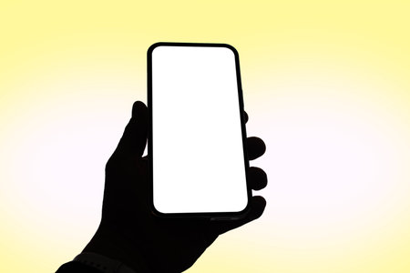 Silhouette of a hand holding a modern smartphone with a blank white screen, isolated on a soft yellow gradient background. Mockup for mobile applications and UI UX design presentationsの写真素材