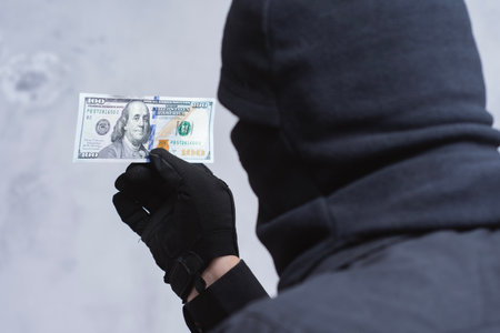 A mysterious figure dressed in a black mask and gloves holds a US hundred dollar bill. Concept bribery, financial crimes, money laundering and corruptionの写真素材
