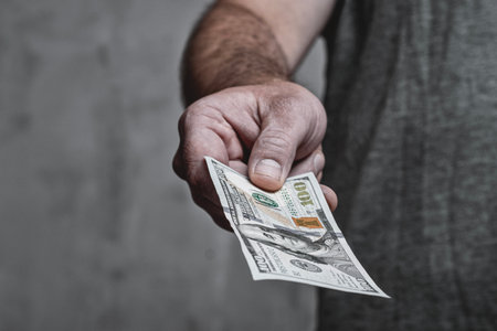 Close-up of a man's hand offering a US one hundred dollar bill. The image represents a financial transaction, payment, bribery, or personal dealの写真素材