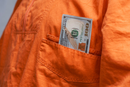 Crisp one hundred dollar bill sticking out of the pocket of an orange jumpsuit. Concept related to corruption, bribery and crimeの写真素材